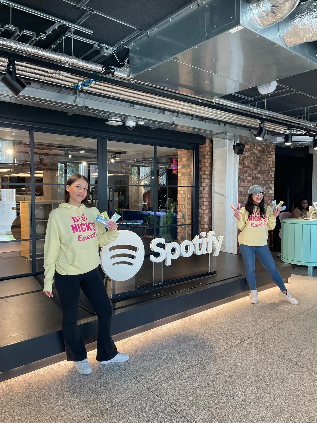 Two FOTS ambassadors in Big Nick's Energy hoodies holding protein bars in front of the Spotify logo in the Spotify office