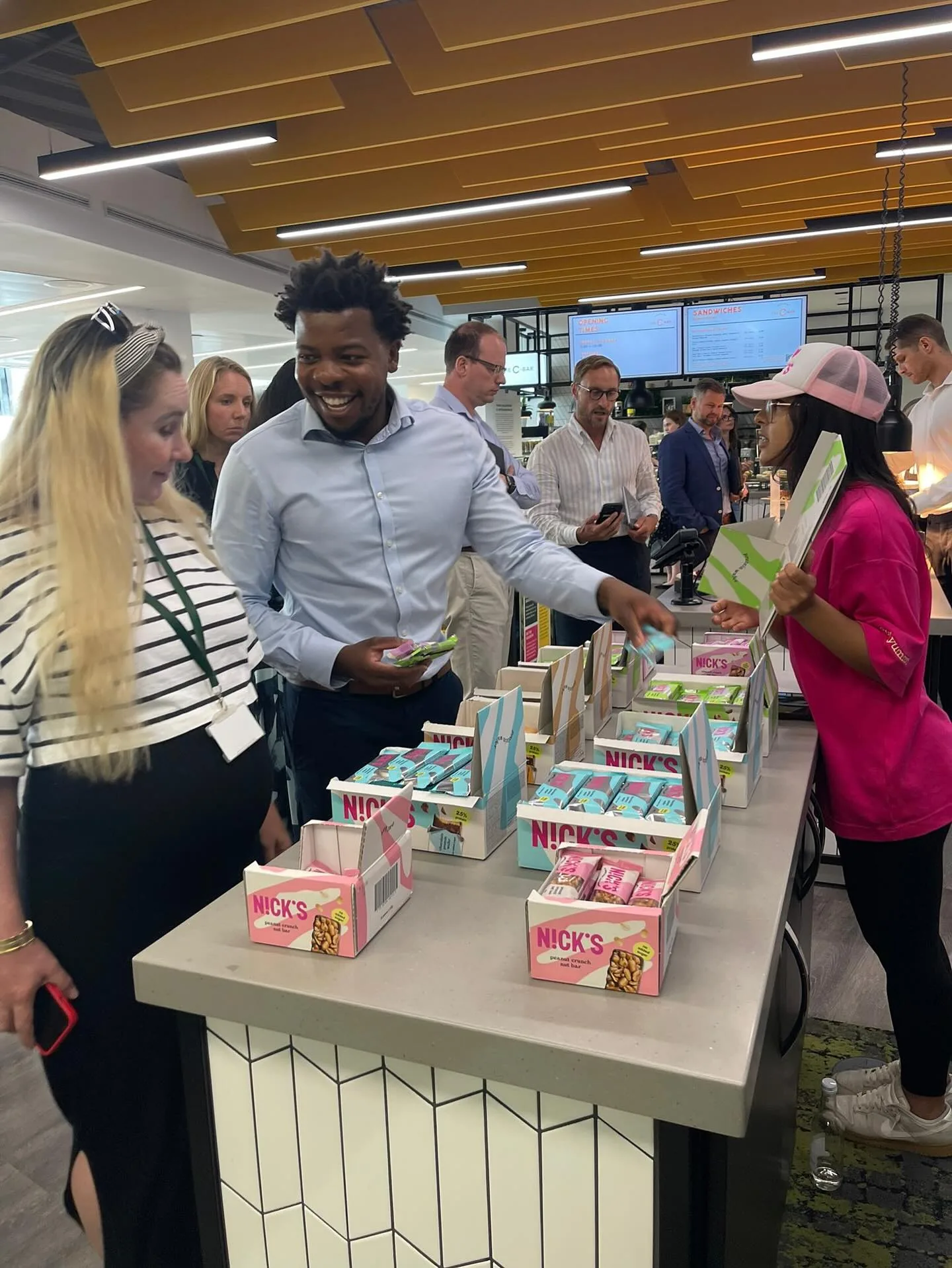 Office workers picking out Nick's protein bars from display boxes at a workplace café sampling table