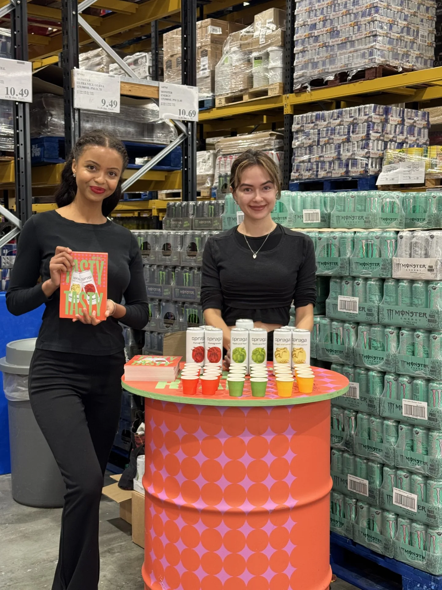 Two FOTS ambassadors at a Spraga tasting barrel with cans and sample cups in a wholesale warehouse